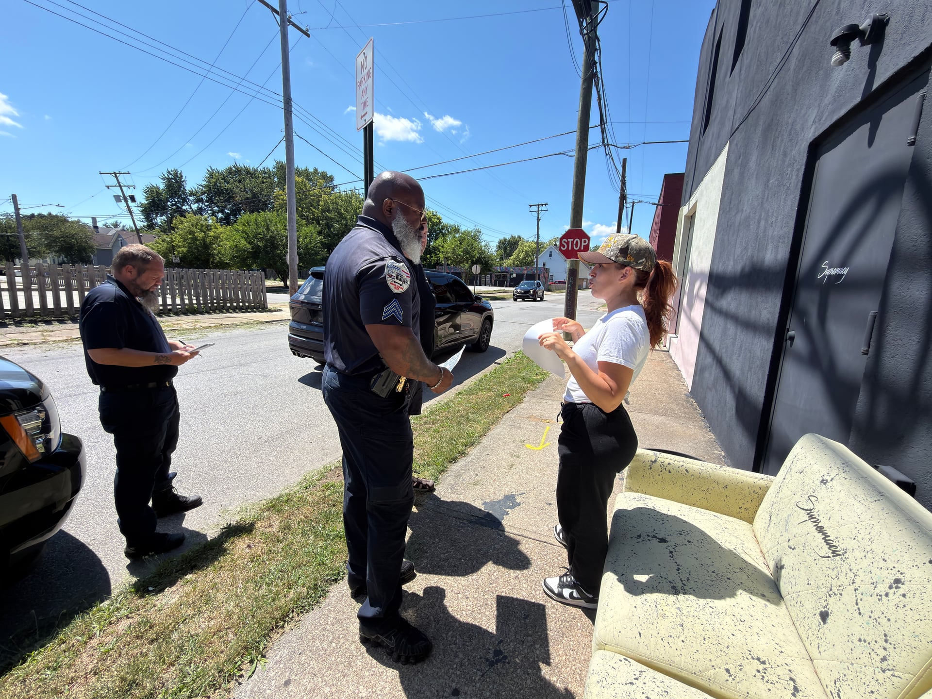 A person holding papers speaking with two people in dark uniforms on a sidewalk.