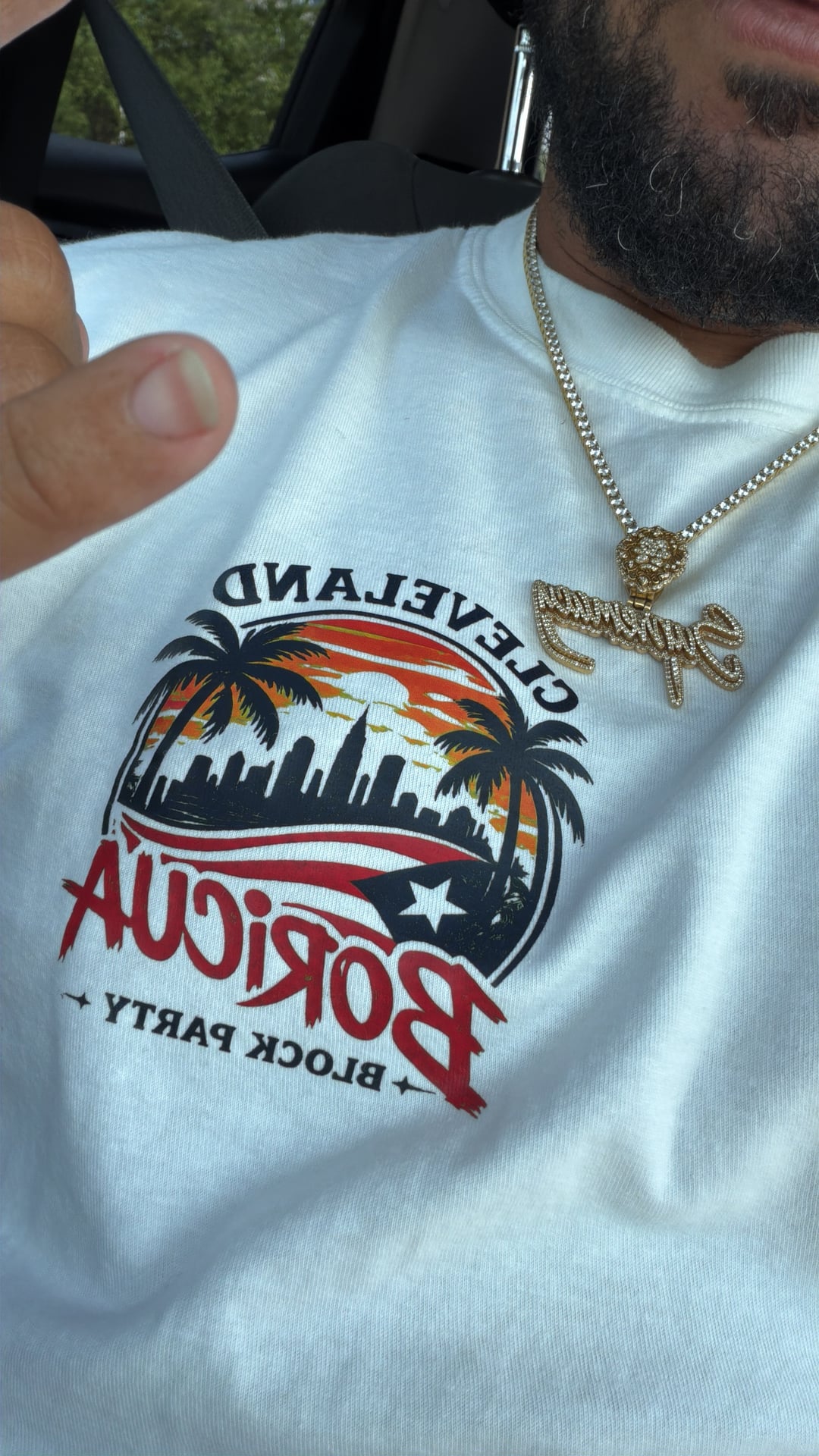 Close-up of a white Cleveland Boricua Block Party shirt and gold necklaces inside a vehicle.