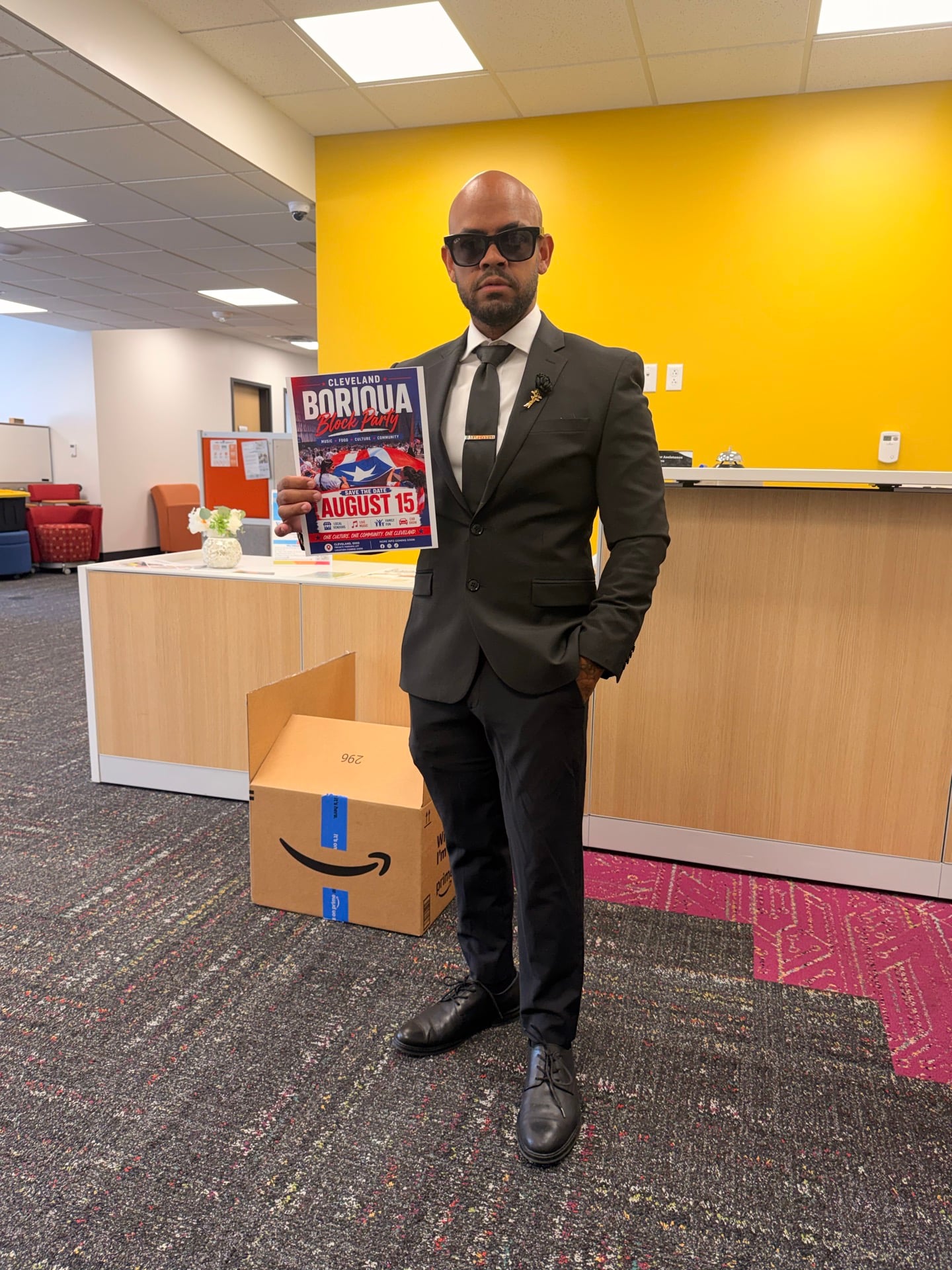 A person in a suit holding an early August 15 block party flyer beside an indoor reception counter.