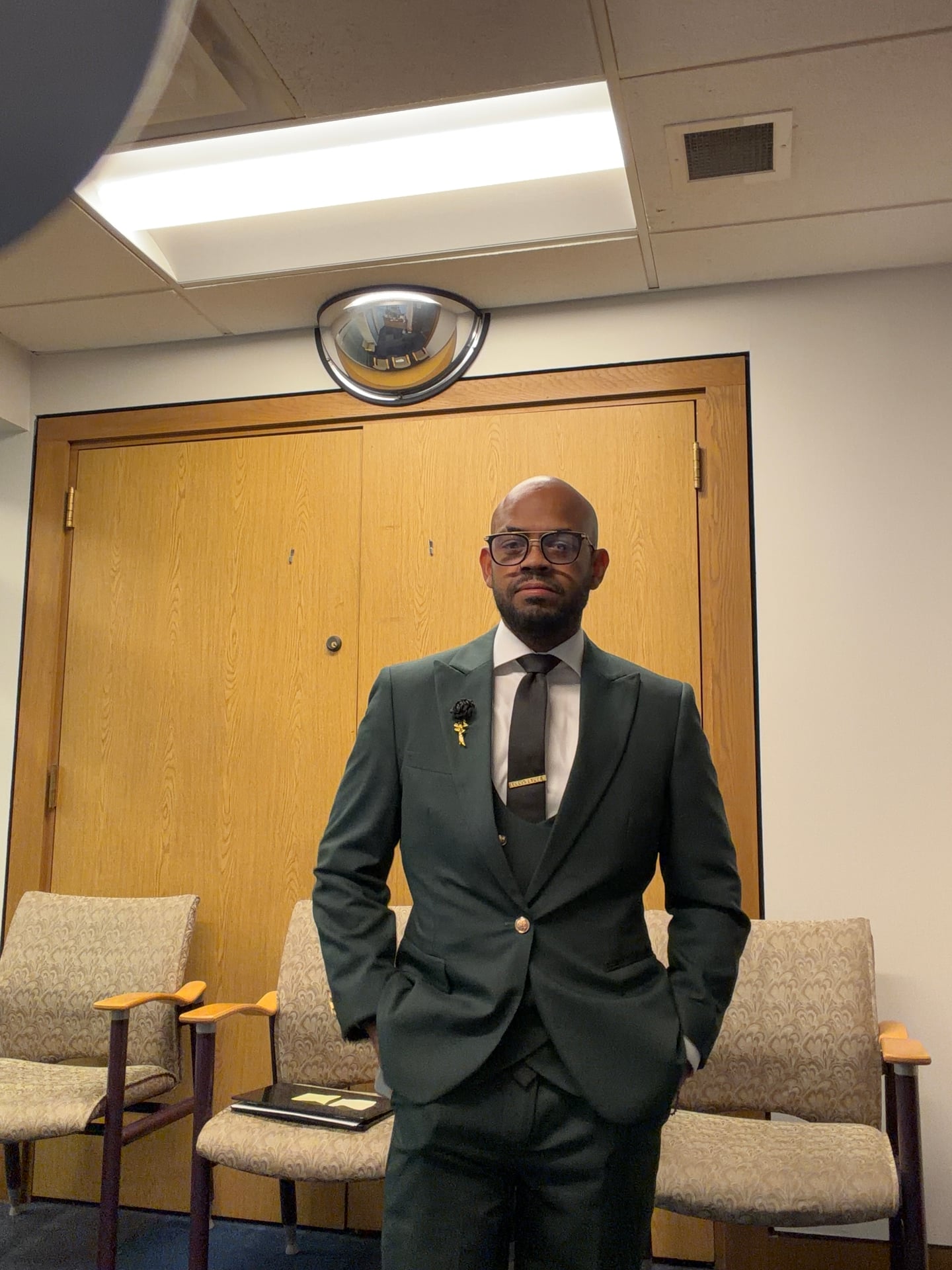 A person in a green suit standing in a room with wooden doors and upholstered chairs.