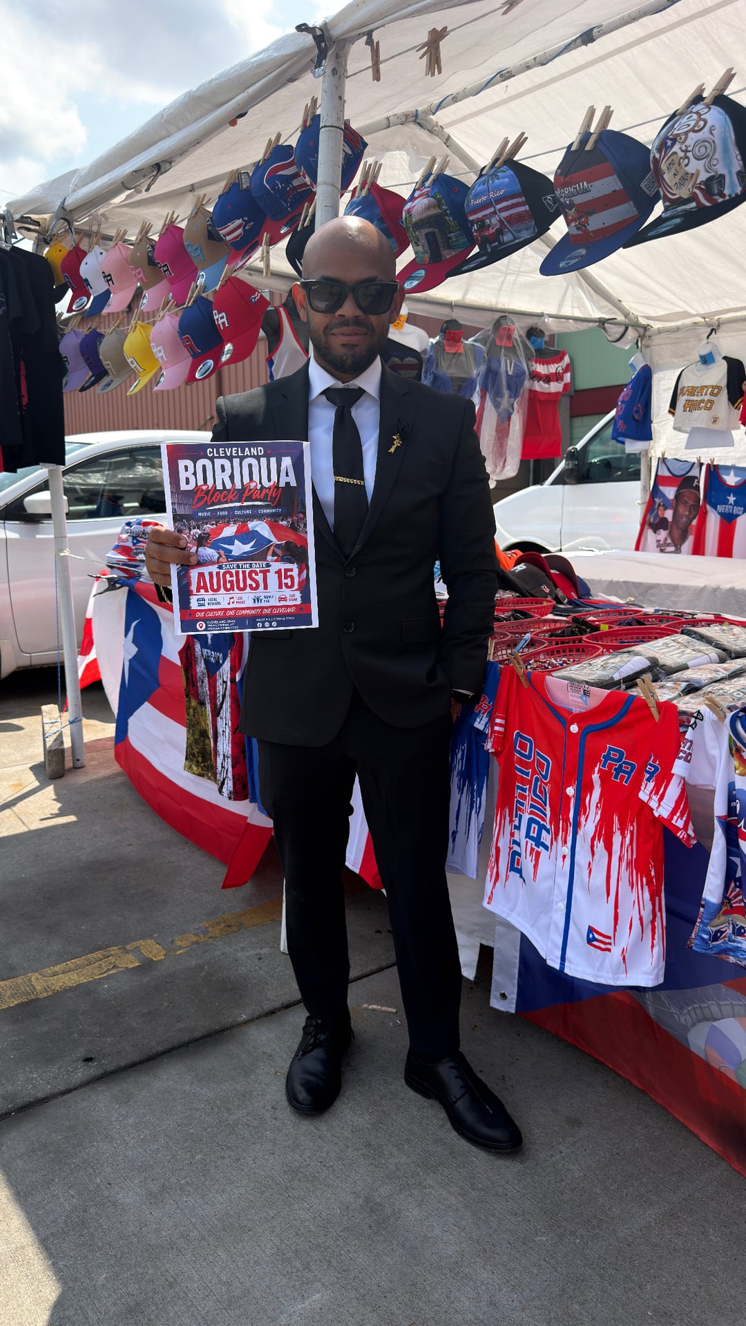 A person holding an early August 15 block party flyer at a stall displaying Puerto Rican-themed hats and clothing.