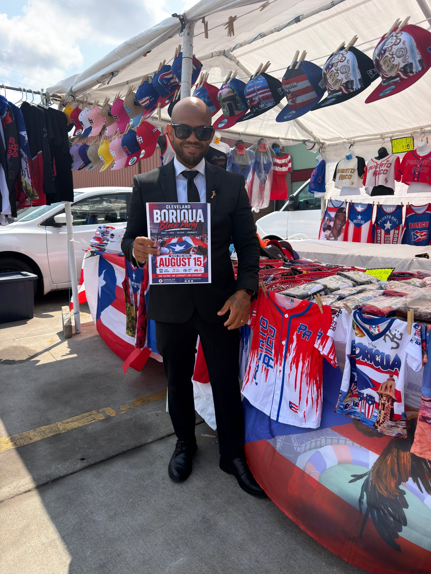 A smiling person in a suit holding an early August 15 block party flyer beneath a canopy of colorful hats.