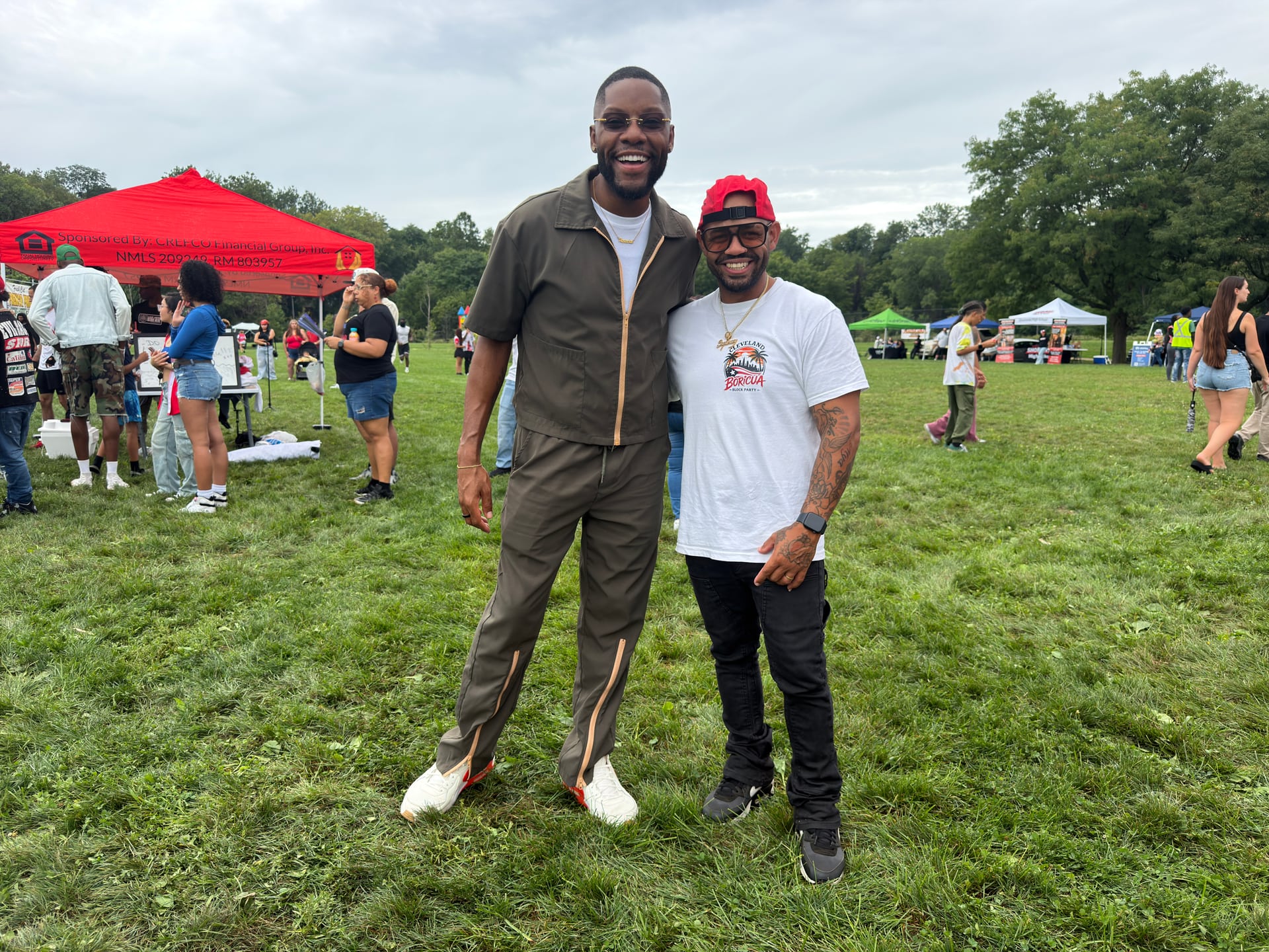 Two people smiling for a photo on the grass with vendor tents behind them.