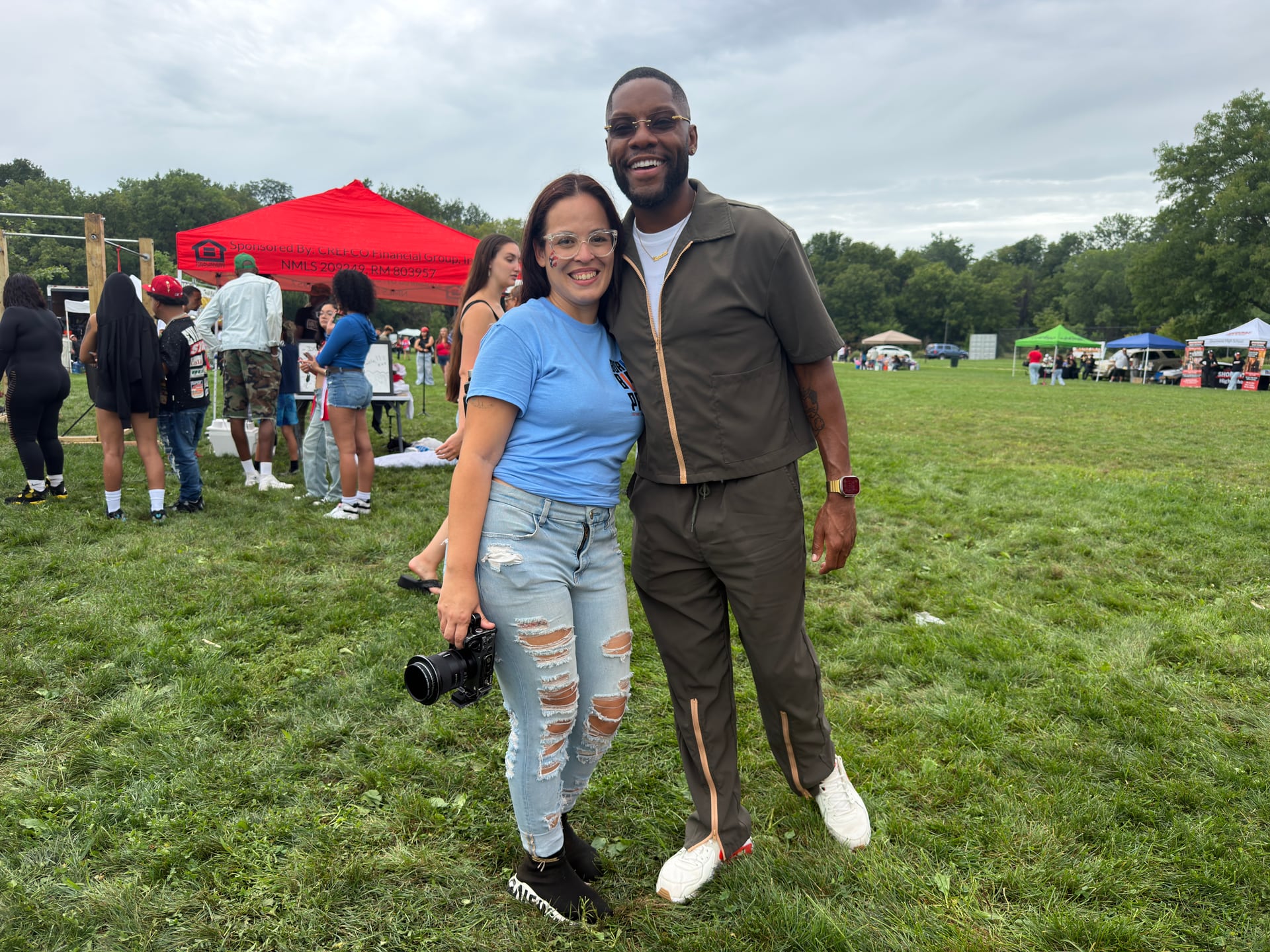 Two people posing on the field, one holding a camera, with colorful vendor tents in the background.