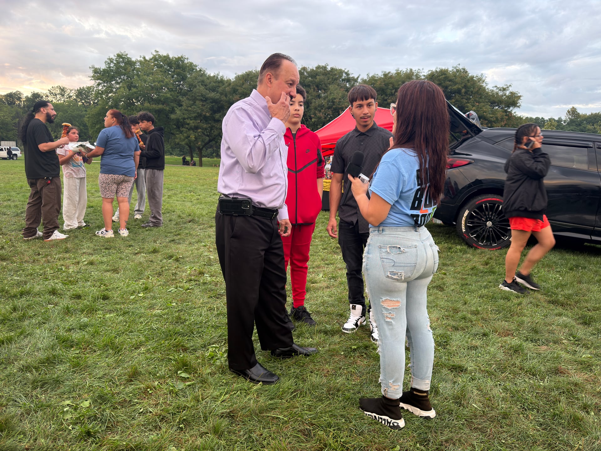 A person holding a microphone talking with a small group on the grass beside a vehicle.