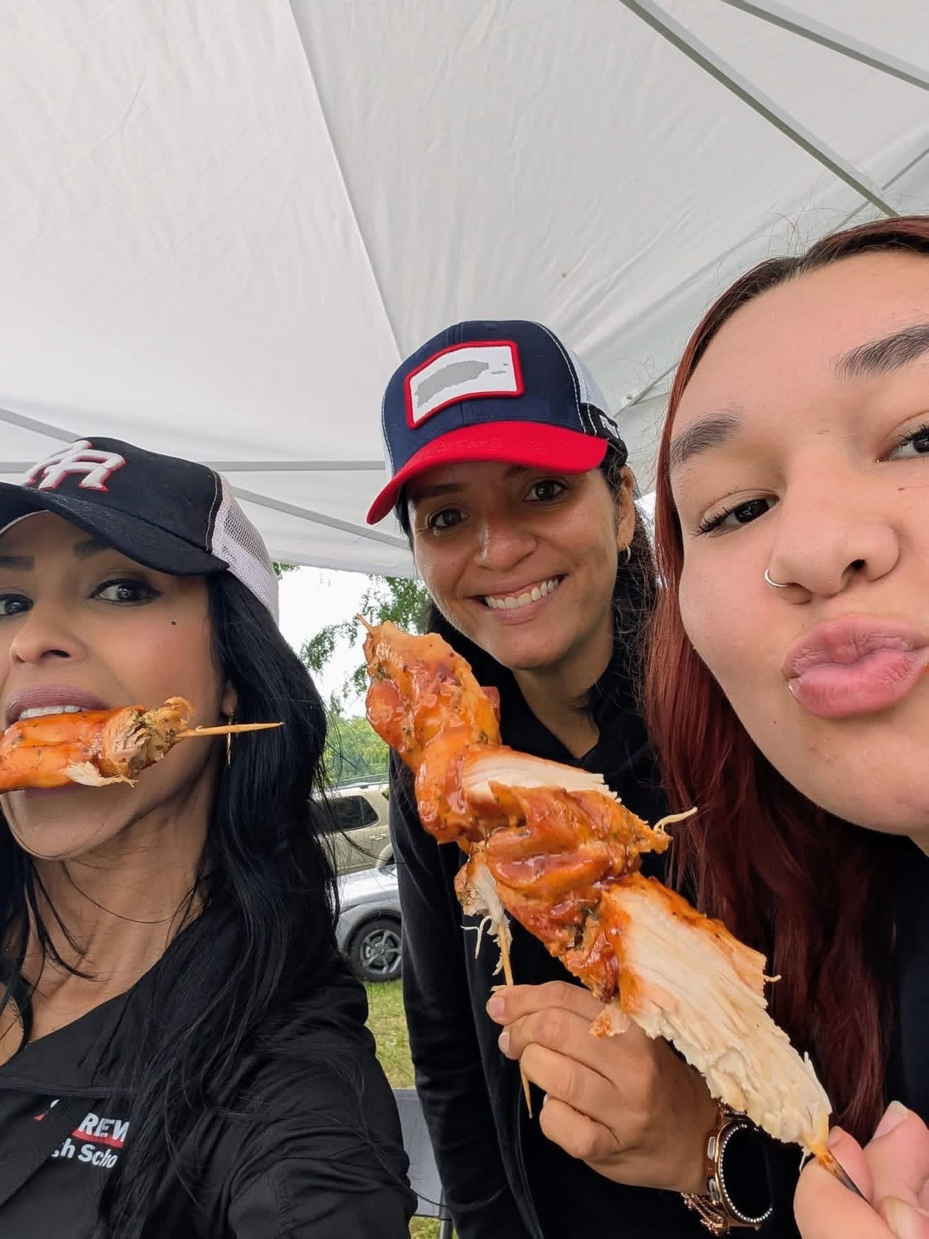 Three people taking a selfie while holding grilled food on skewers beneath a canopy.