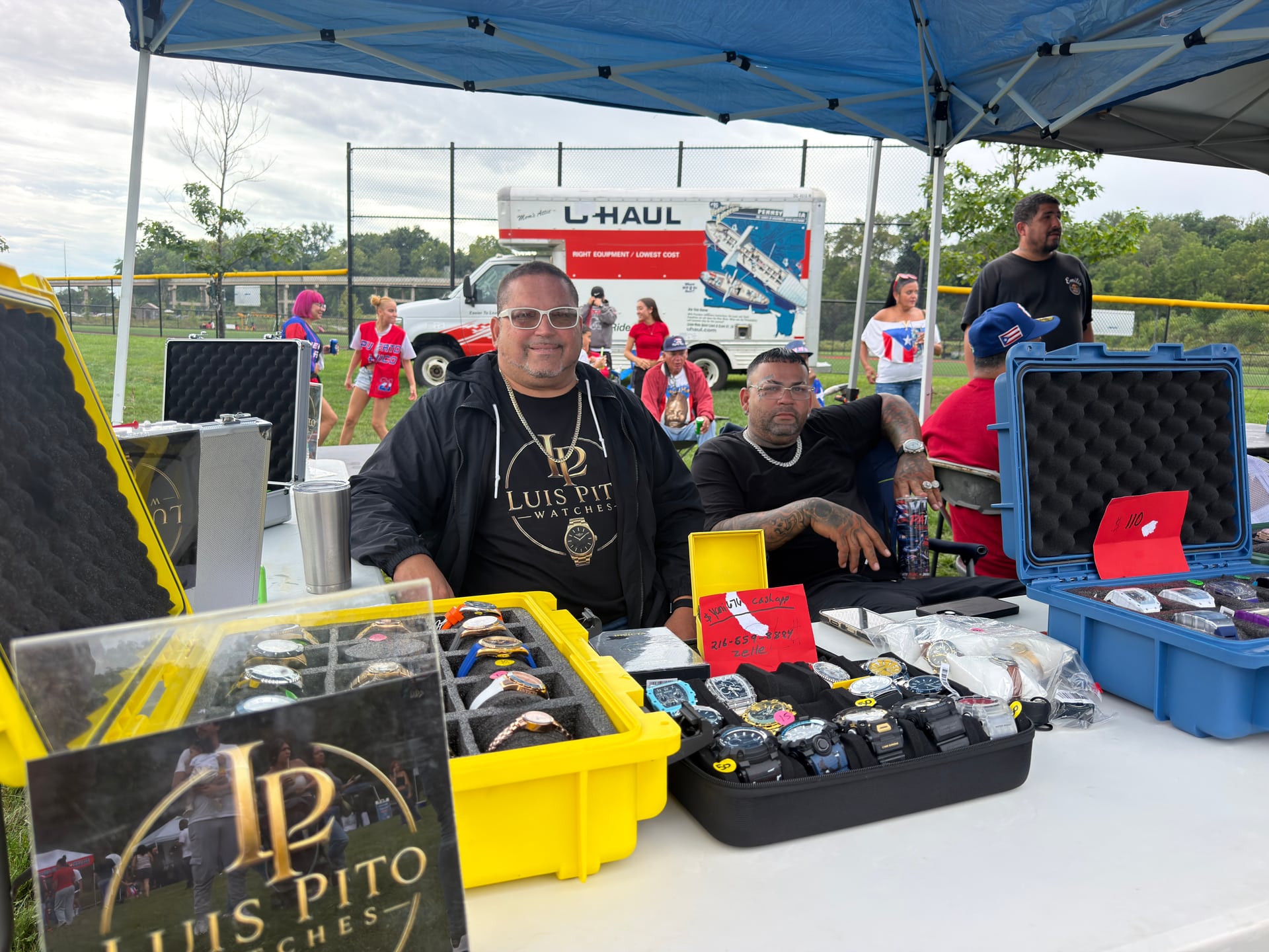 People at a watch vendor table with wristwatches displayed in open cases.