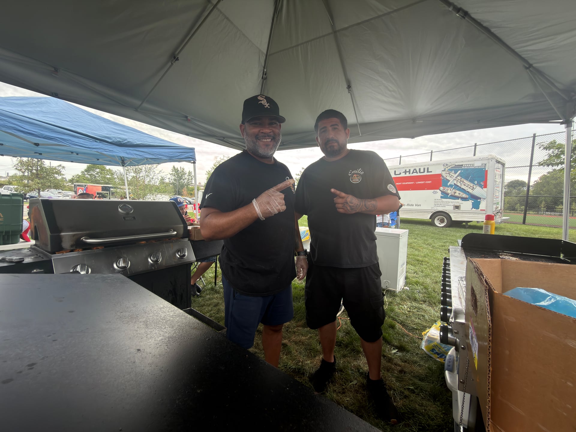 Two people posing beside grills and cooking equipment underneath a vendor canopy.