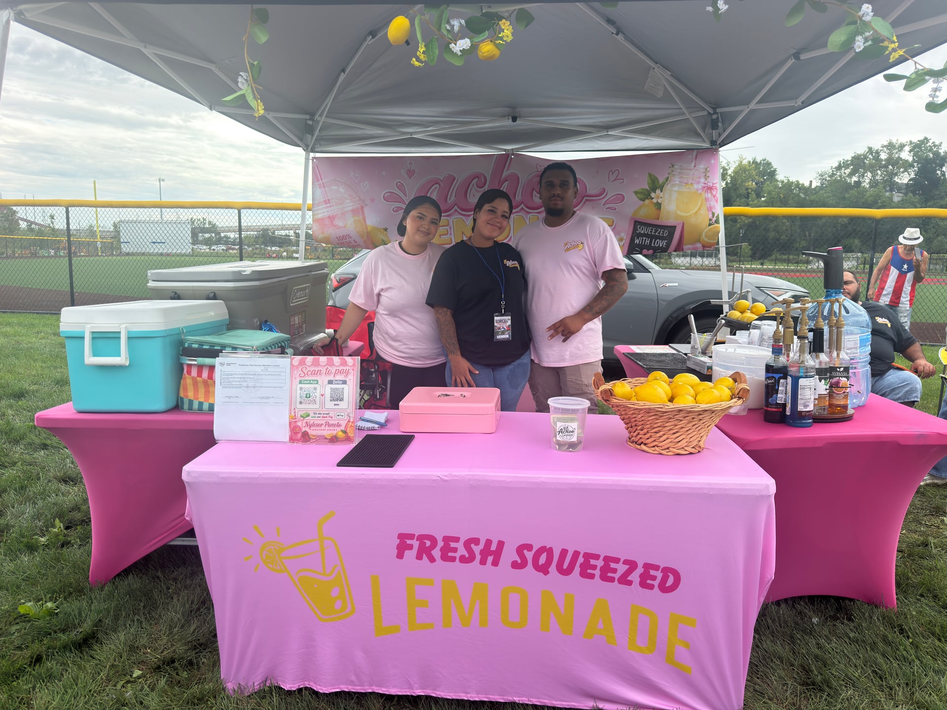 Three people at a bright pink lemonade stand with lemons, coolers, and flavored syrups.