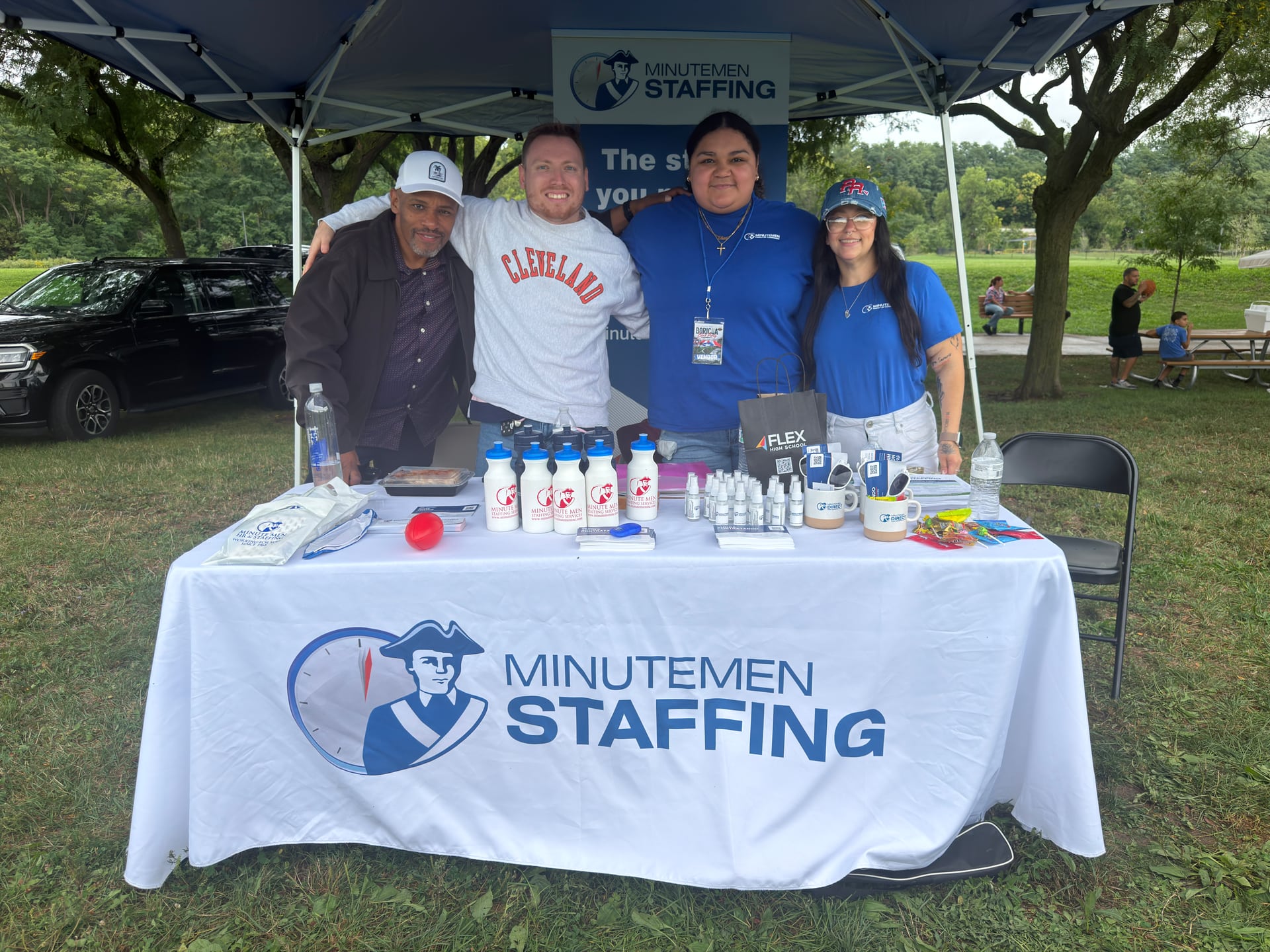 Four people posing together behind a Minutemen Staffing table with promotional items.