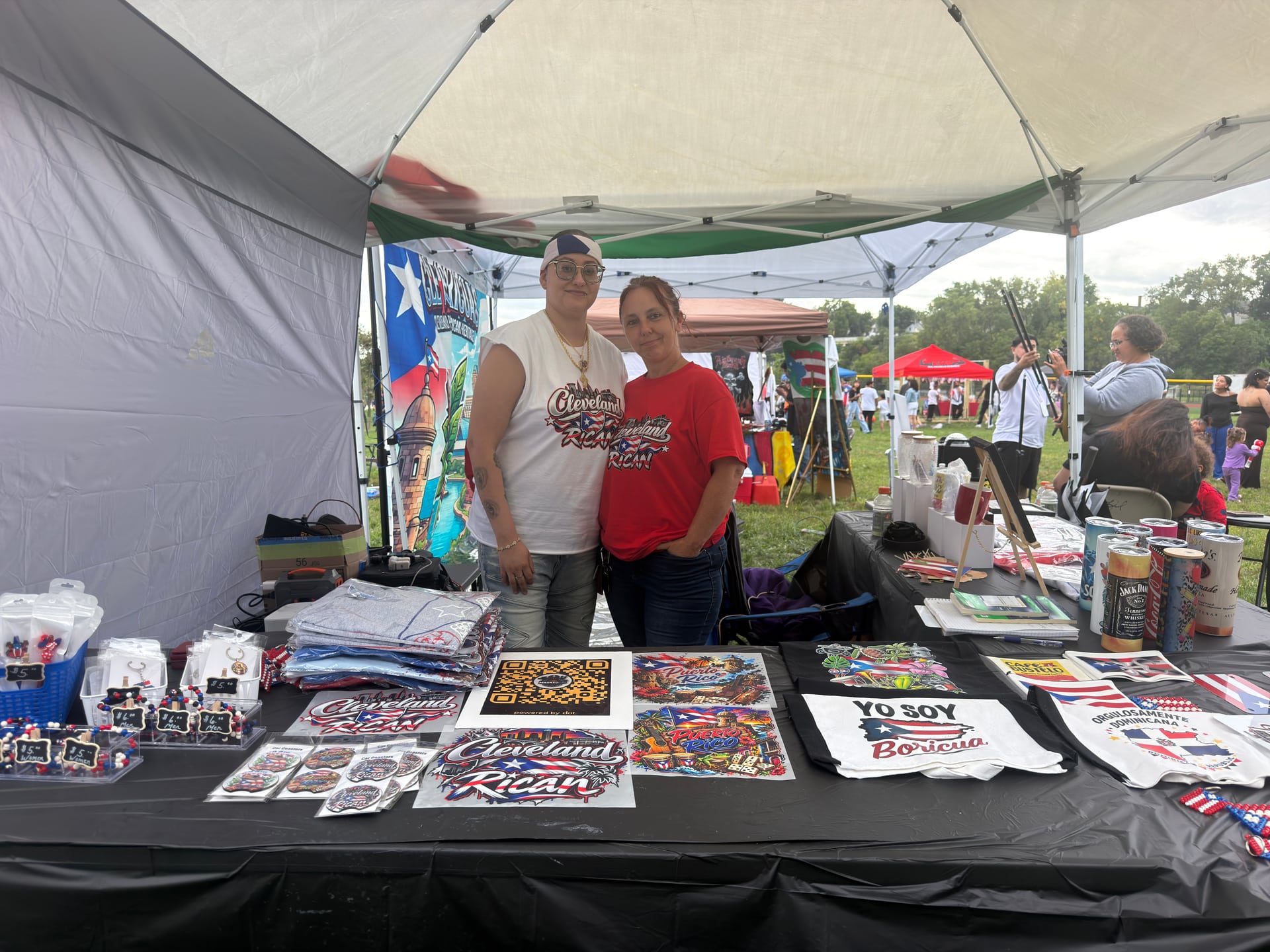 Two people at a vendor stall displaying Puerto Rican-themed shirts, bags, and other merchandise.