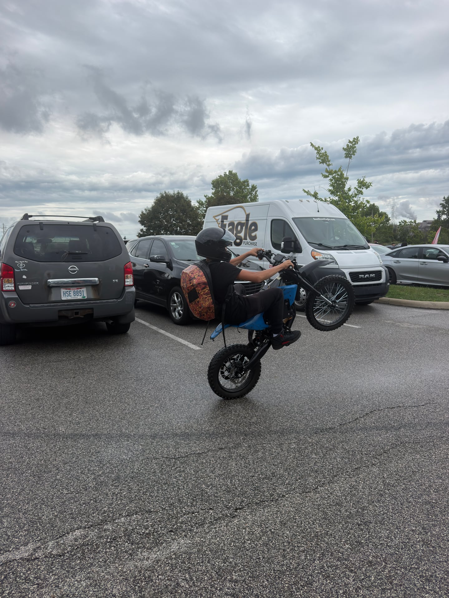 A helmeted rider balancing a motorbike on its rear wheel in a parking area.