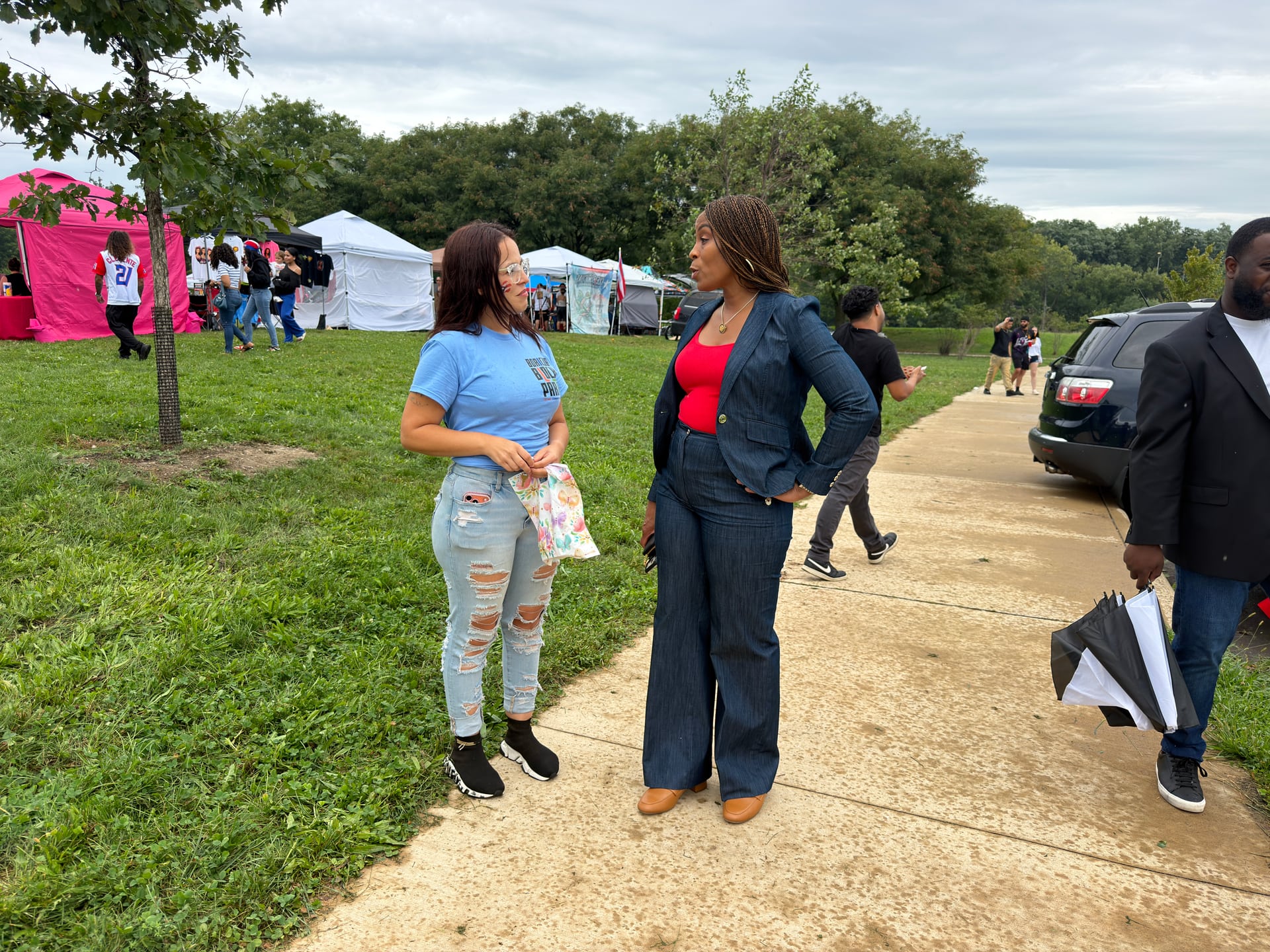Two people talking on a park path with tents and other visitors in the background.