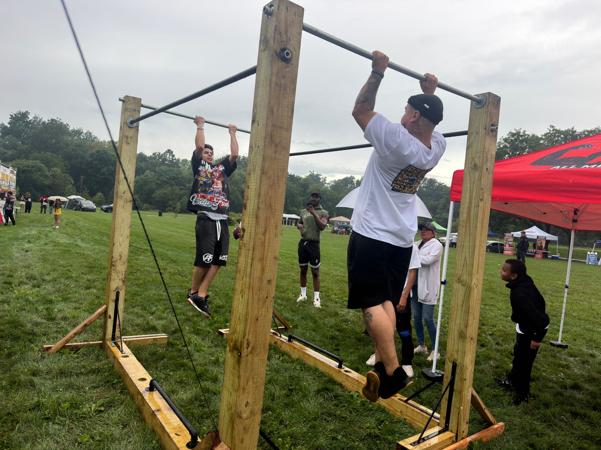 Two people using outdoor pull-up bars while others look on from the grass.