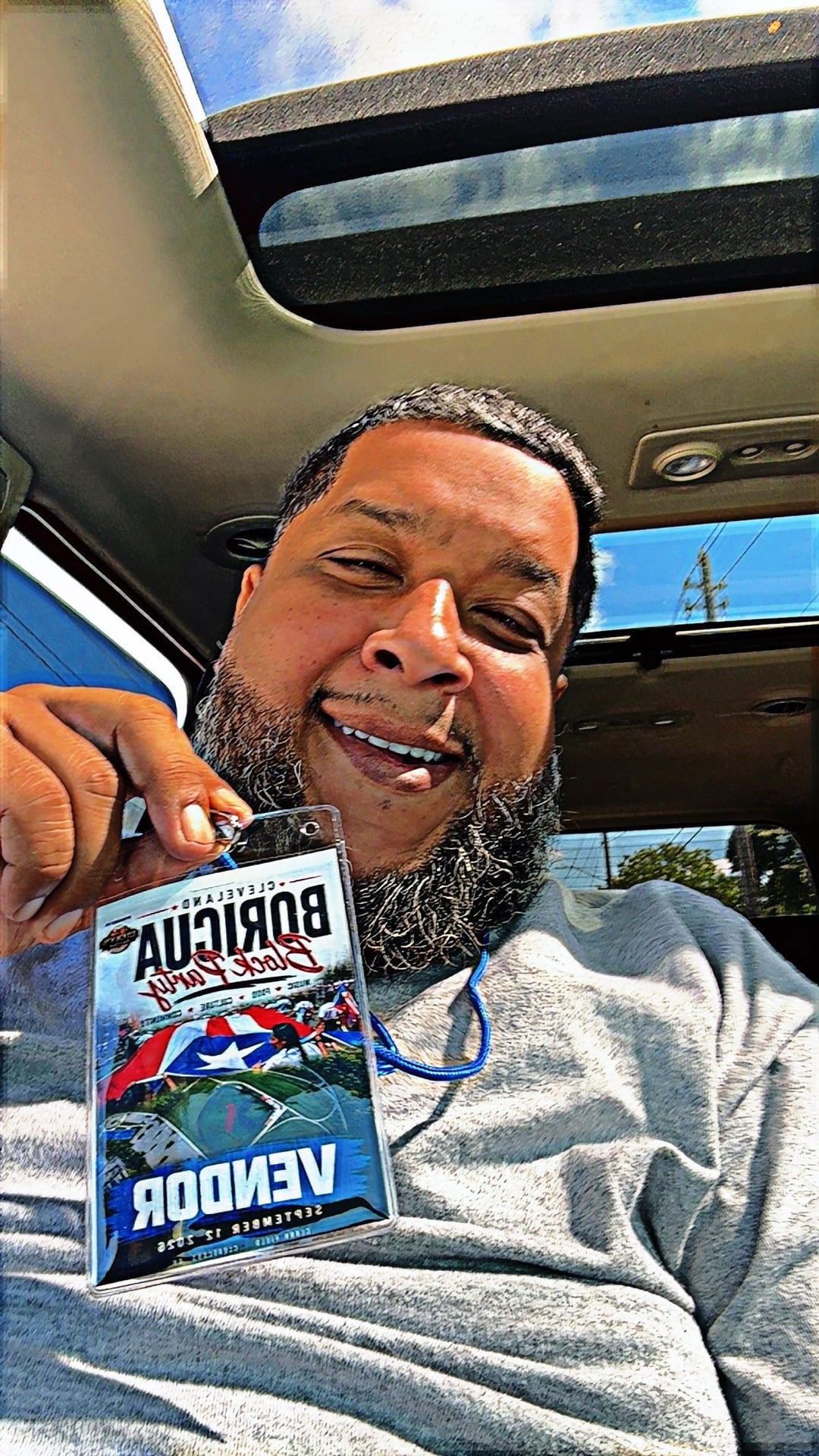A smiling person holding a block party vendor badge while sitting inside a vehicle.