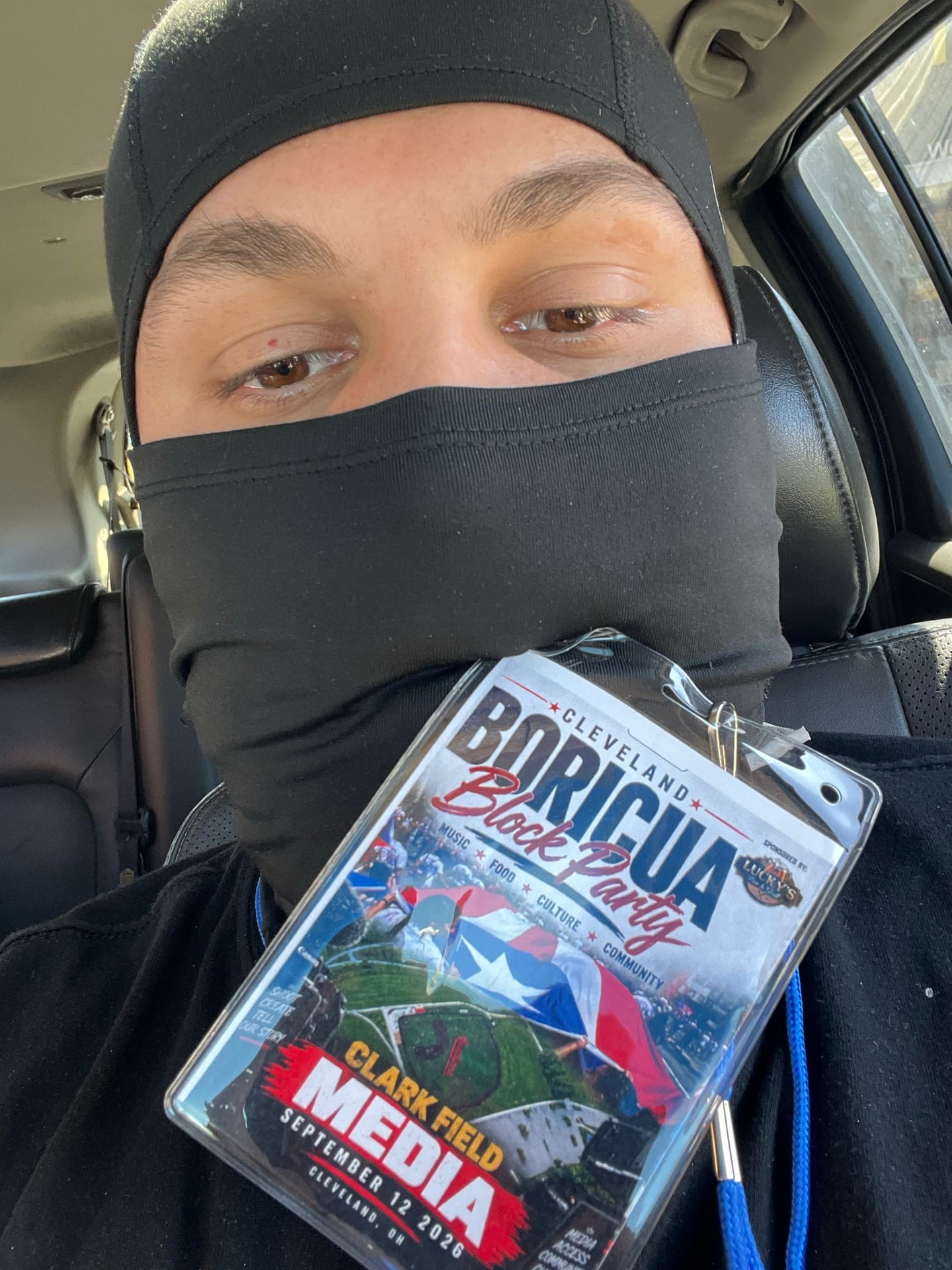 A person wearing a black face covering and holding a block party media badge inside a vehicle.